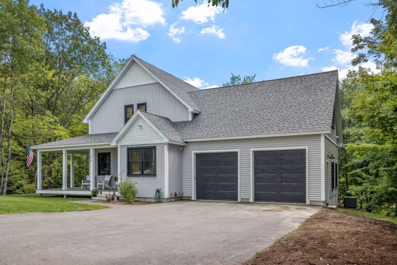 Two-story house with gray siding, a covered front porch, and an attached two-car garage. Enhanced by quality craftsmanship and exterior cleaning, this home stands beautifully against a backdrop of trees and a clear sky.