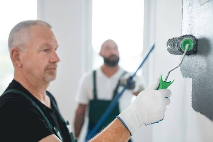 A man paints a wall with a roller, showcasing quality craftsmanship, while another man stands in the background holding a pole; both wear work overalls during this home remodeling project.