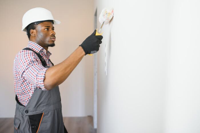A man wearing a hard hat and overalls uses a paint roller to paint a white wall, showcasing professional painting skills ideal for Knoxville home improvement projects.