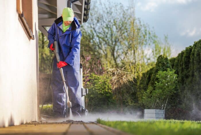 A person in protective clothing uses a pressure washer for exterior cleaning of an outdoor patio beside a house, surrounded by greenery, enhancing Knoxville home improvement.